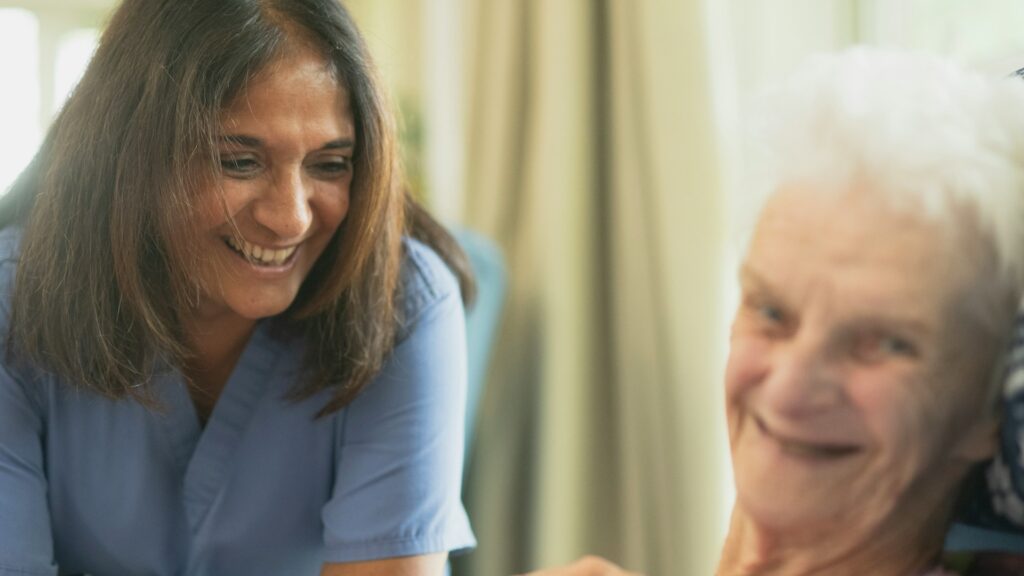 Zorgverlener lacht terwijl ze contact maakt met oudere vrouw in thuiszorgsituatie – thuisgerichte WLZ-zorg